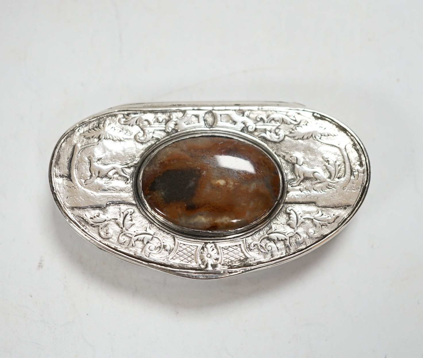 An early 19th century Scottish? white metal oval snuff box, with banded agate base and inset agate lid, decorated with dogs and scrolls, 69mm.